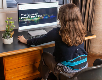 Load image into Gallery viewer, Person sitting at a desk using a computer while wearing a DuoTherm with 'The Future of Physics Medicine' displayed on the screen.
